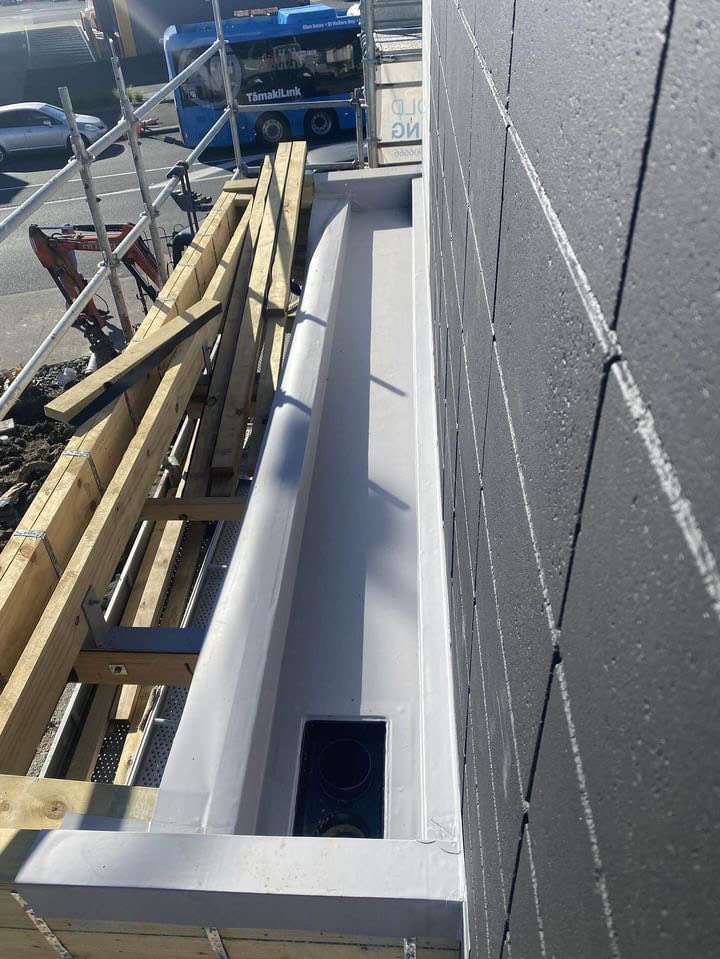 Grey membrane box gutter with a square outlet, next to a block wall and scaffolding